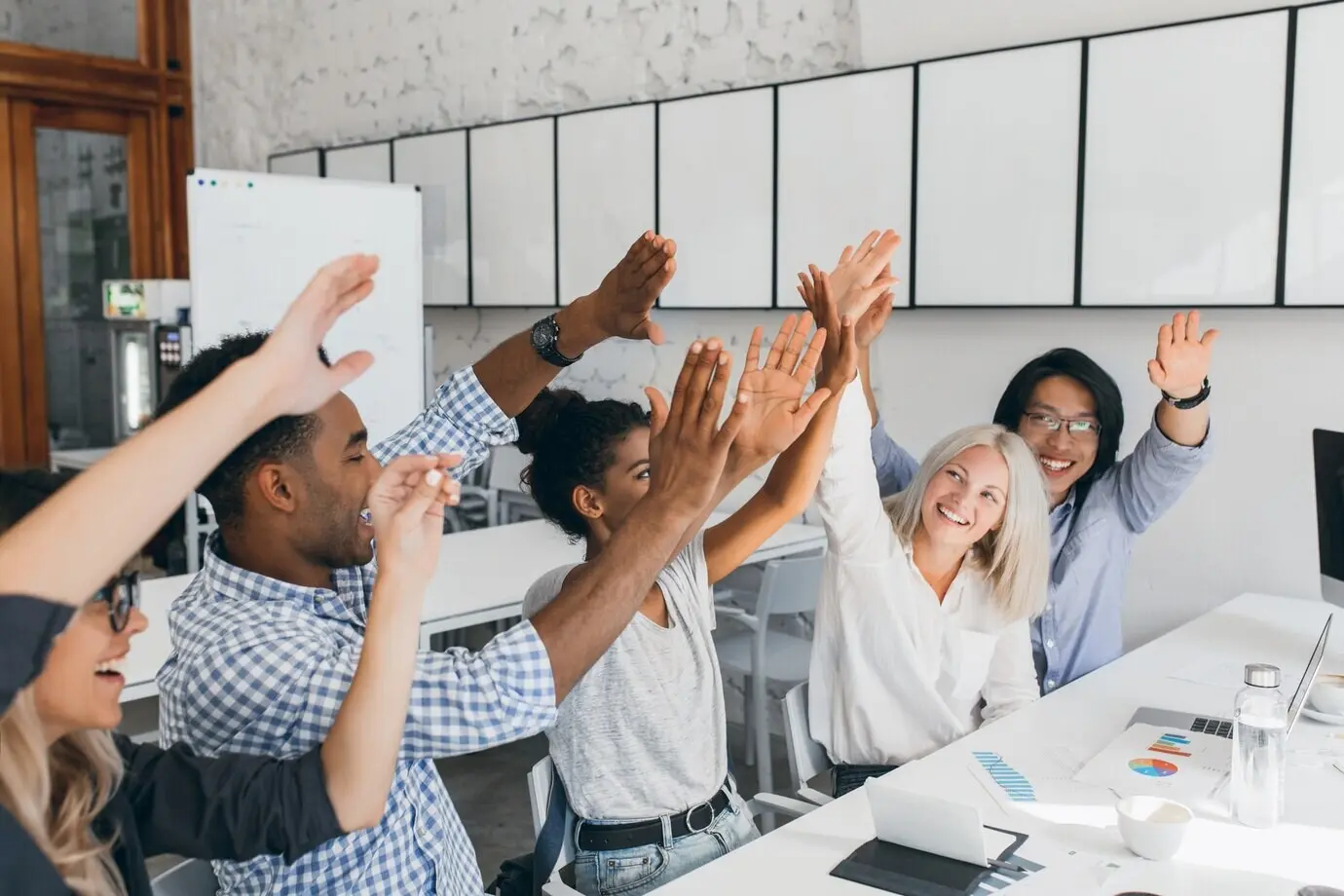 An adorable woman with a short blonde haircut high-fives her African female coworker. Indoor photo of joyful colleagues celebrating the start of vacation at the workplace.