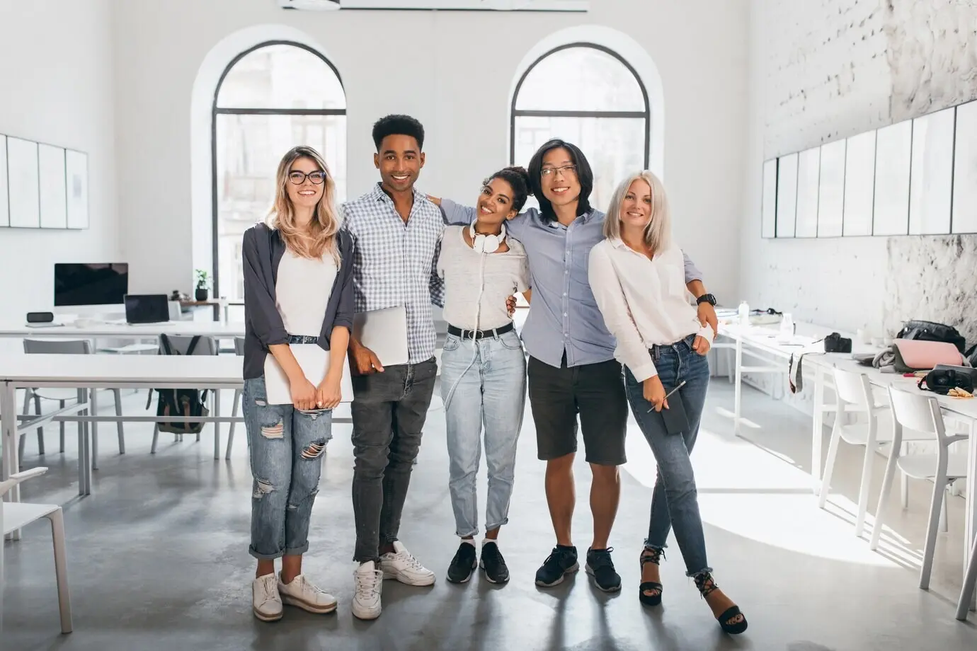 Full-length portrait of a shy blonde woman in white sneakers holding a laptop after a seminar, standing beside an African friend. Excited international students posing together after a lecture in a spacious hall.
